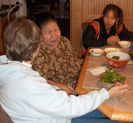 Nanta, Ann and Dona are sitting at a kitchen table.  Dona is eating a salad, Nanta's and Anns' dishes are empty.  Nanta is leaning over to say something to Dona.